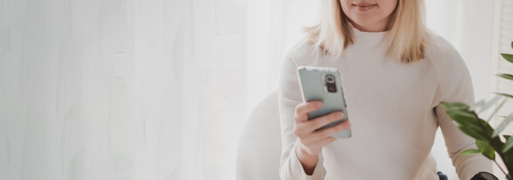 A person in a light-colored sweater is holding a smartphone, sitting near a window with sheer curtains and next to a potted plant.