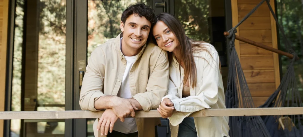 A man and woman lean over a table, smiling and engaged in an activity. An easel with a floral painting is in the background.