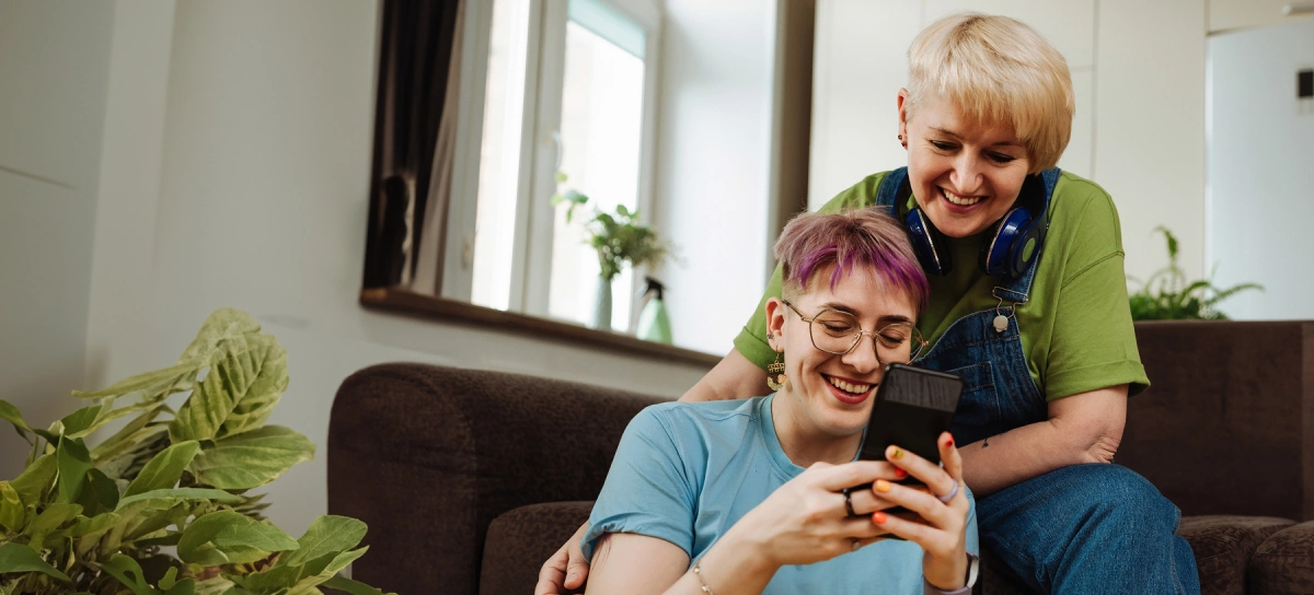 Two people sit on a couch looking at a smartphone screen and smiling. One person has short blonde hair and headphones around their neck, while the other has purple hair and glasses. Indoor plants are visible.