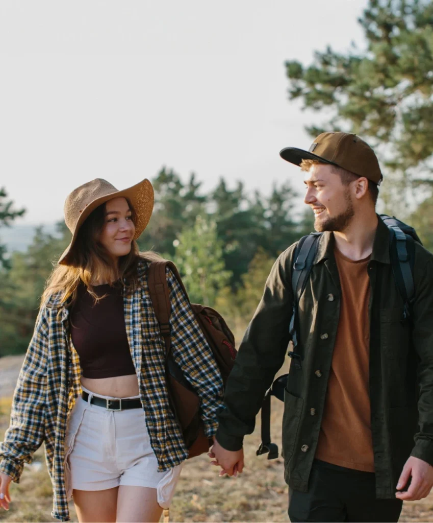 A couple holding hands, wearing casual outdoors clothing and hats, walking on a trail with trees in the background. Both are carrying backpacks.