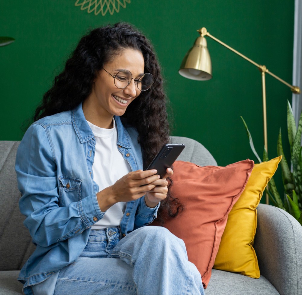A woman wearing glasses and a denim jacket sits on a sofa, smiling while looking at her smartphone. She is surrounded by colorful cushions and a green wall with a lamp in the background.