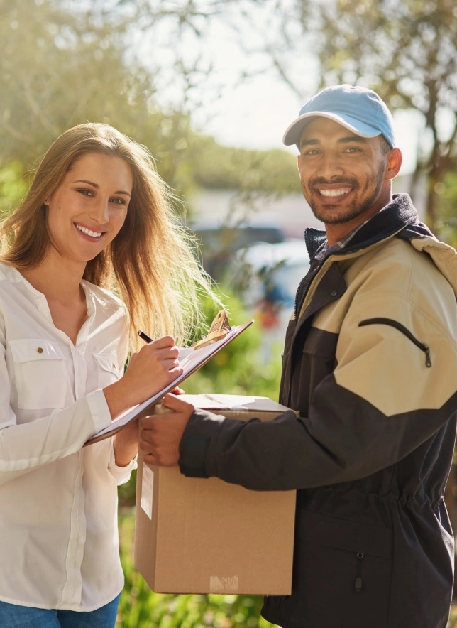 A smiling woman signs a clipboard while a smiling delivery person holds a package outdoors.
