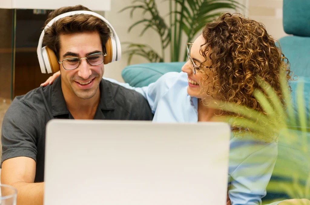 A man wearing headphones and a woman sitting next to him smile at a laptop screen.