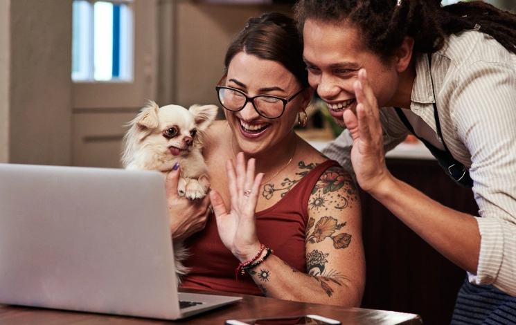 Two people smiling and looking at a laptop screen. One is wearing headphones, and the other has their arm on the other's shoulder. They are sitting in a bright room with plants in the background.