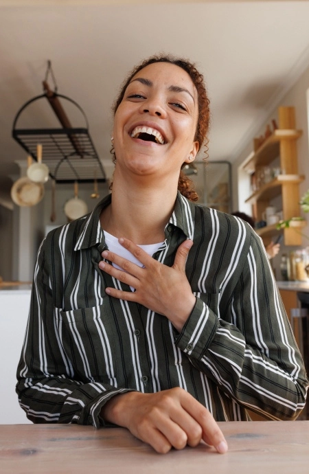 A person wearing a striped shirt is laughing with a hand on their chest while sitting at a table in a kitchen.