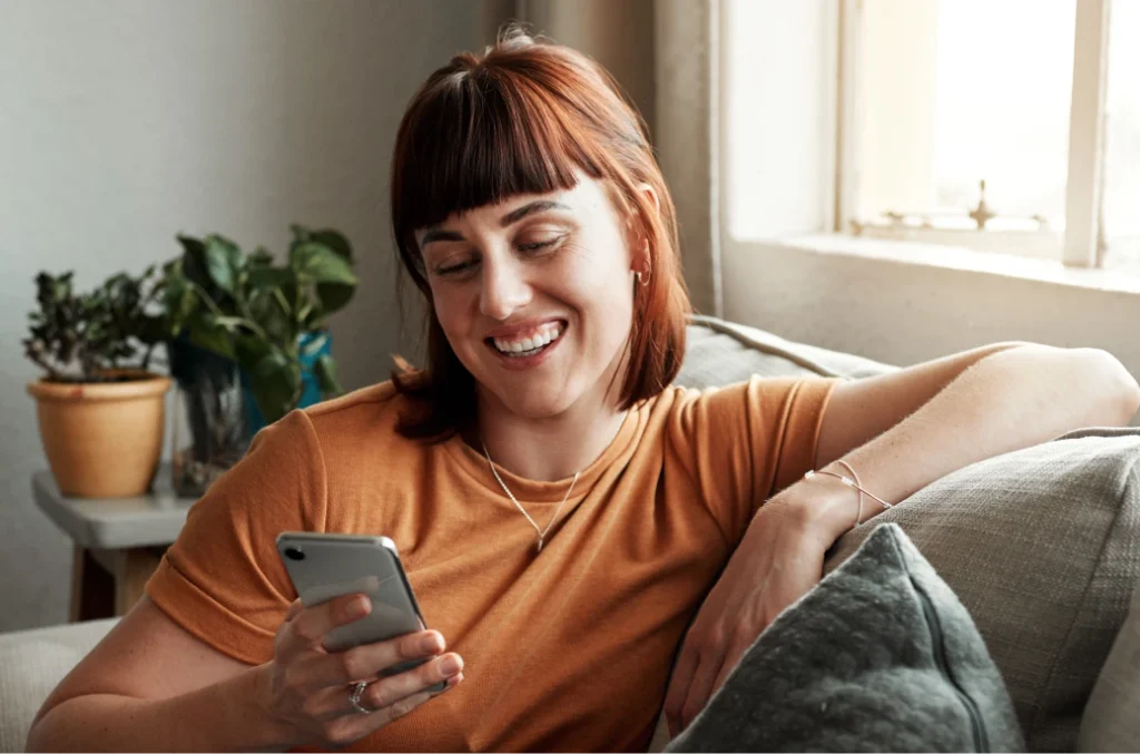 A woman with auburn hair and a fringe smiles while looking at her phone, sitting on a couch next to a plant.